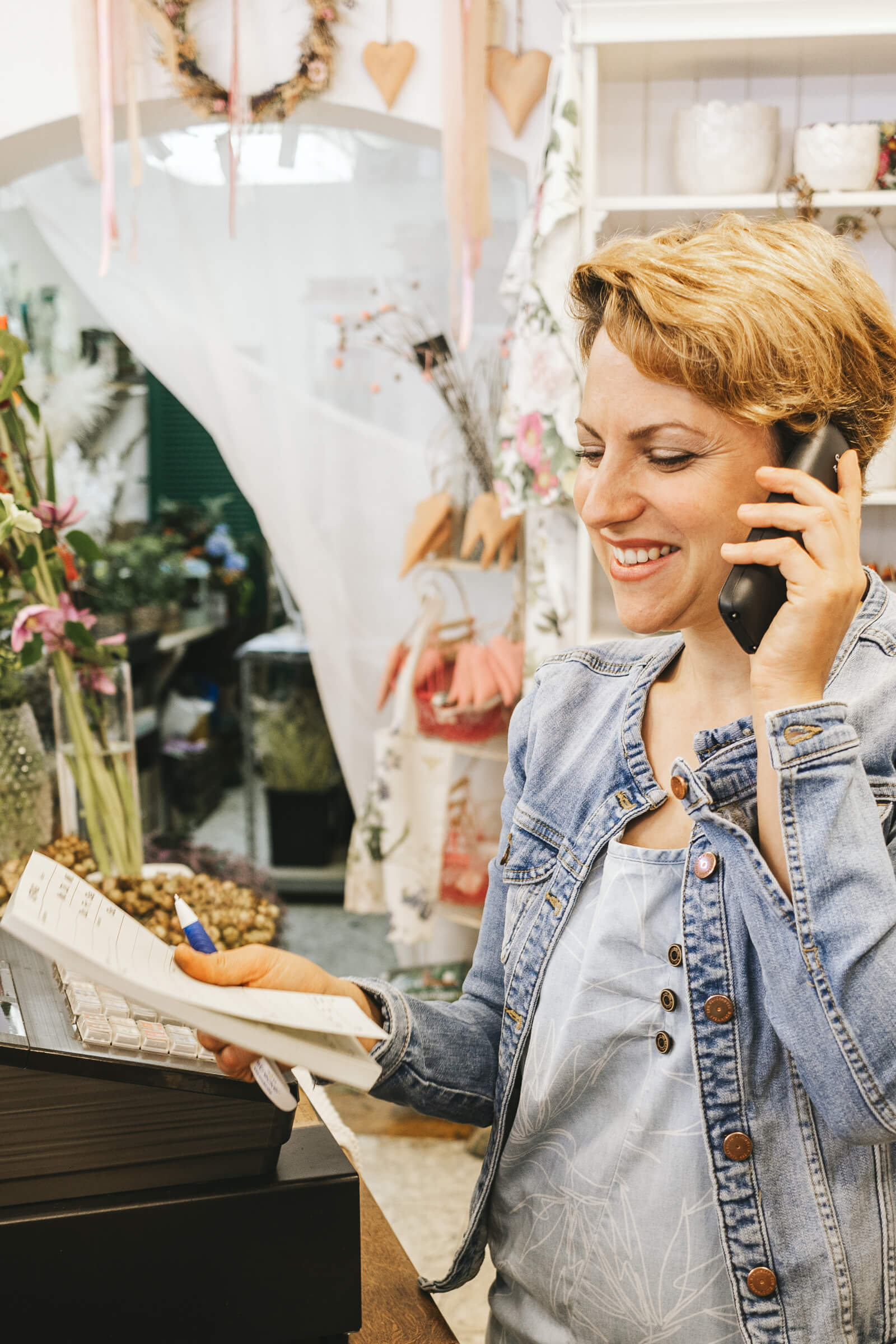 Eine Frau in einer Jeansjacke telefoniert und hält einen Notizblock in der Hand, während sie an der Kasse eines Blumenladens steht.