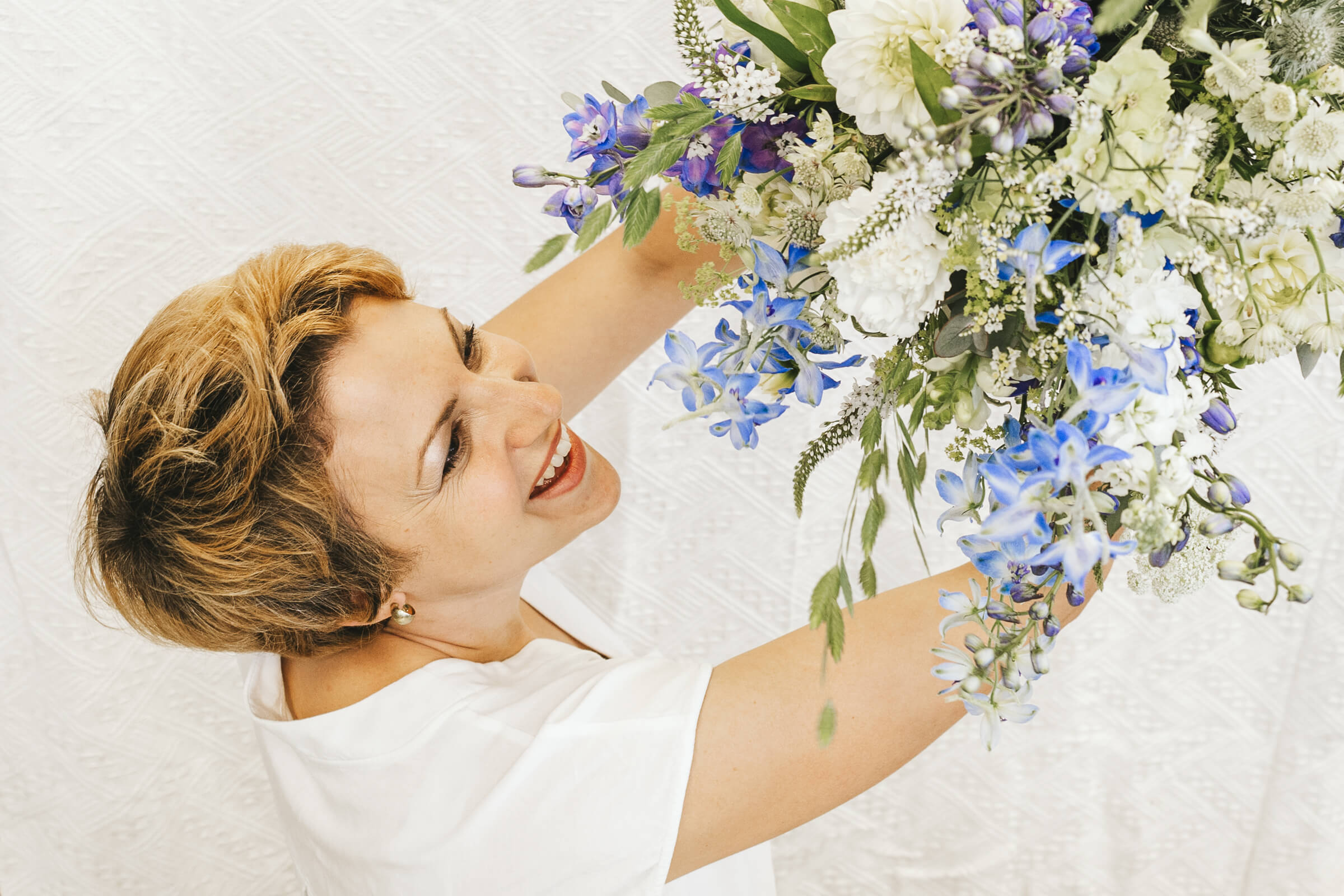 Eine Frau mit kurzen Haaren hält einen üppigen Strauß aus blauen und weißen Blumen vor einem strukturierten weißen Hintergrund.